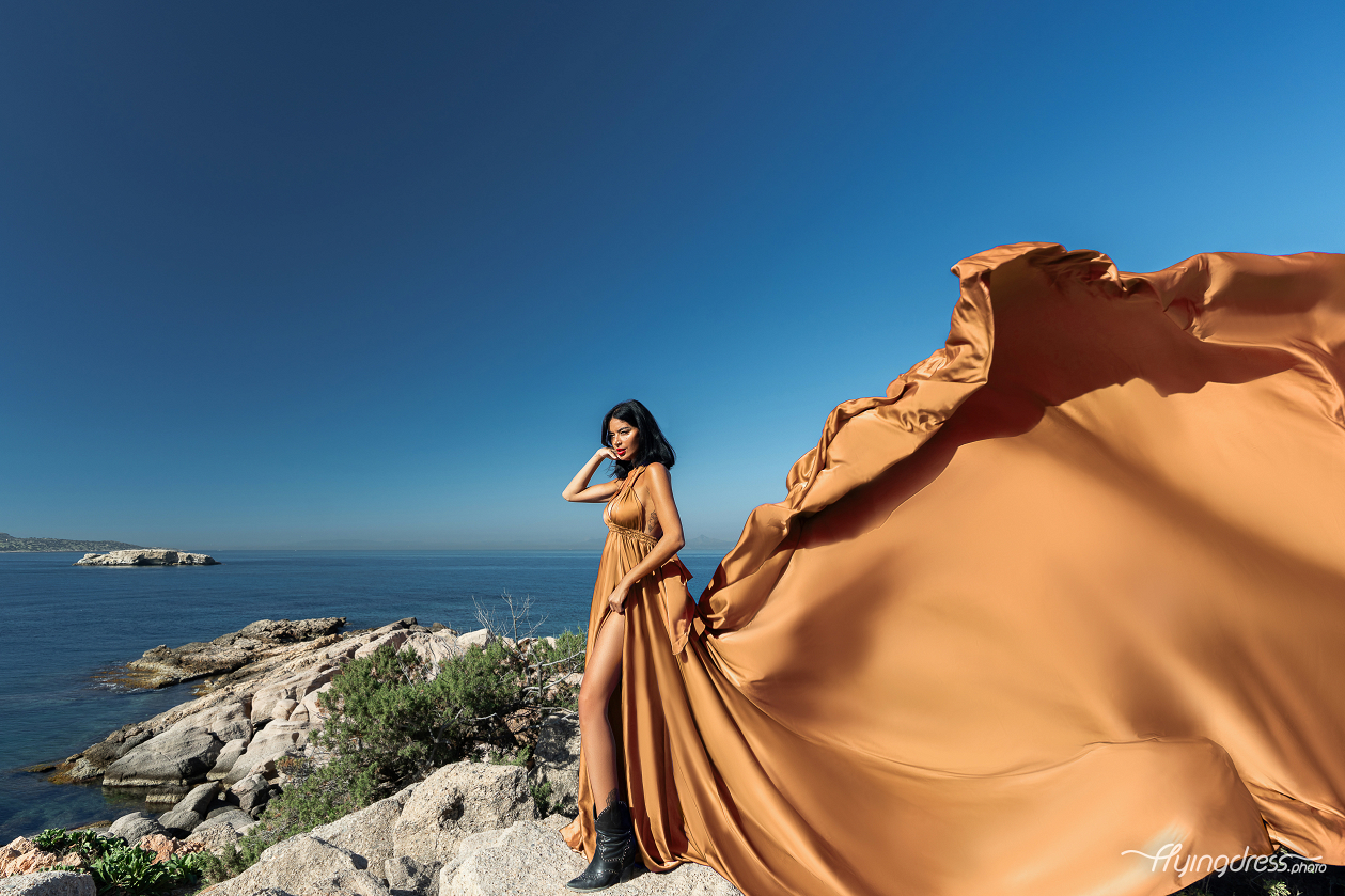 A woman in a flowing golden dress and black boots stands on a rocky cliff, with the tranquil blue waters and coastal scenery of Kavouri, Athens extending behind her.